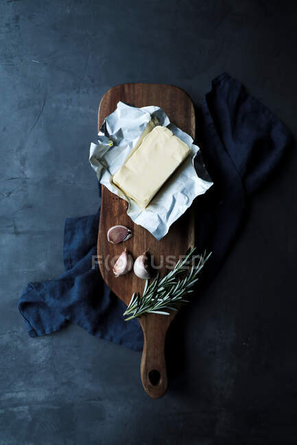 Vista superior del paquete abierto de mantequilla en composición con dientes de ajo y tallos de romero sobre tabla de cortar de madera y textil colocado sobre mesa oscura en cocina de estilo rústico - foto de stock