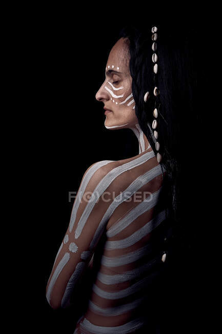 Side view of naked tender beautiful brunette Native American woman with white striped painted on body covering breast standing in dark on black background — Stock Photo