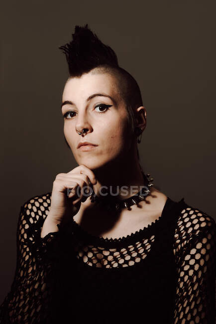 Serious adult lady with mohawk touching chin and looking at camera while standing against brown background — Stock Photo