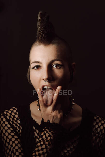 Feminino punk saindo da língua entre dois dedos e olhando para a câmera contra o fundo preto — Fotografia de Stock