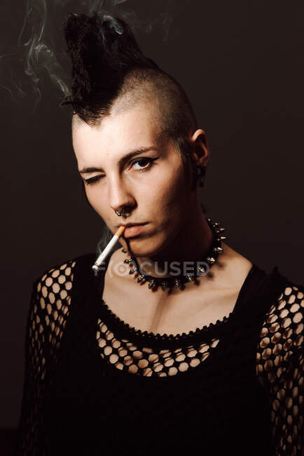 Adult female with mohawk and piercing looking at camera and igniting cigarette with lighter on black background — Stock Photo