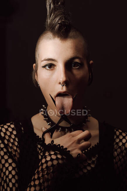 Mujer punk sobresaliendo lengua entre y mirando a la cámara sobre fondo negro - foto de stock