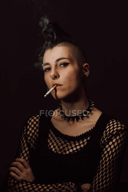 Adult female with mohawk and piercing looking at camera and igniting cigarette with lighter on black background — Stock Photo
