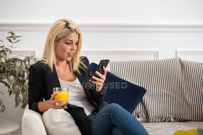 Mujer rubia feliz en ropa casual disfrutando de jugo de naranja fresco y el teléfono inteligente de navegación mientras está sentado en el sofá en casa - foto de stock