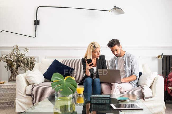 Mujer rubia positiva con taza de jugo de navegación teléfono inteligente y sentado en el sofá cerca de novio étnico escribiendo en el teclado del ordenador portátil en la sala de estar del apartamento moderno - foto de stock