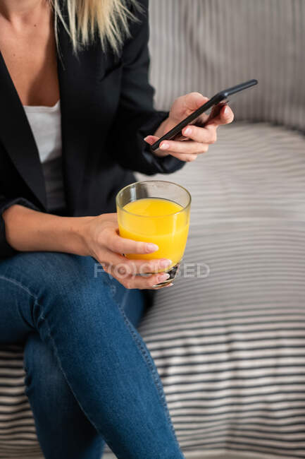 Обрезанная неузнаваемая блондинка в повседневной одежде наслаждаясь свежим апельсиновым соком и просматривая смартфон, сидя дома на диване — стоковое фото