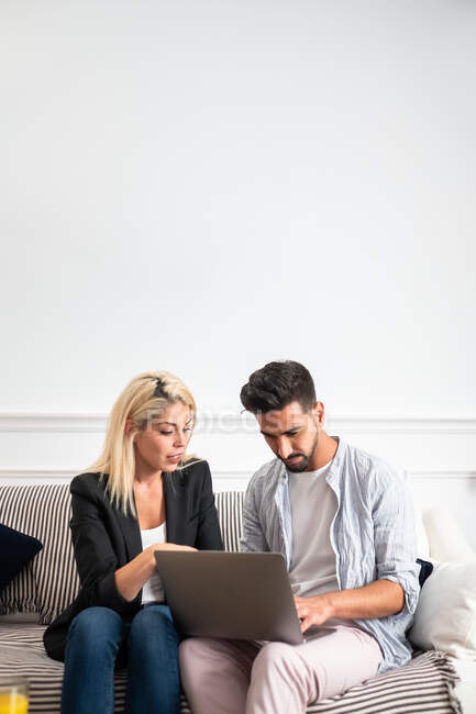 Blonde woman and ethnic bearded handsome man in casual clothes sitting on sofa and browsing laptop together at home — Stock Photo