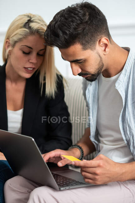 Jovem casal multiétnico sentado no sofá e fazendo compras on-line, juntamente com cartão de crédito em um laptop — Fotografia de Stock