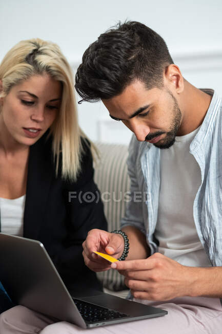 Jeune couple multiethnique assis sur un canapé et effectuant des achats en ligne avec carte de crédit sur un ordinateur portable — Photo de stock