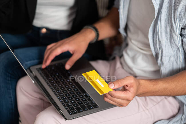 Обрізані невпізнавані руки людини, що сидять на дивані і роблять онлайн-покупки з кредитною карткою на ноутбуці — стокове фото