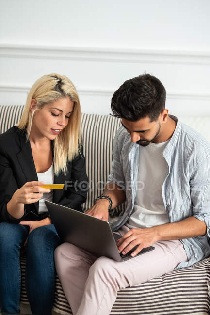 Delighted blonde female reading credit card credentials to ethnic boyfriend with laptop while sitting on sofa and making online purchases together — Stock Photo