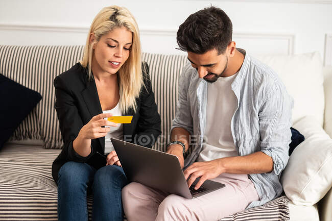 Восхитительная блондинка, улыбающаяся и читающая удостоверения кредитной карты веселому этническому парню с ноутбуком, сидящему на диване и совершающему совместные покупки в интернете — стоковое фото
