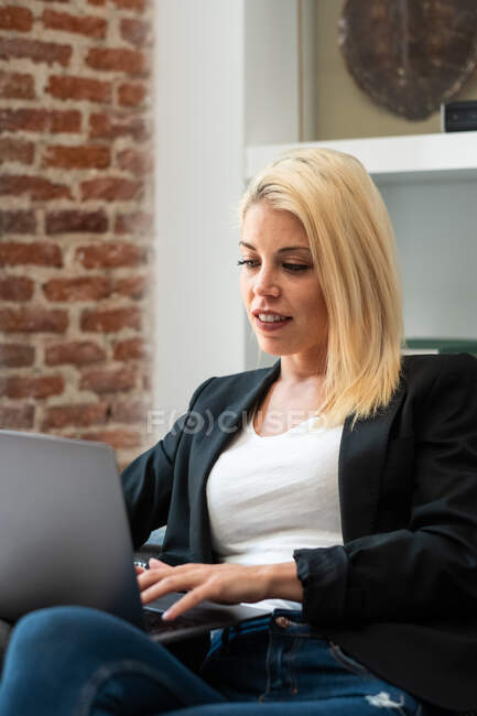 Mulher de negócios loira alegre sorrindo e navegando laptop enquanto sentado em poltrona confortável perto da lareira no quarto acolhedor trabalhando remotamente de casa — Fotografia de Stock
