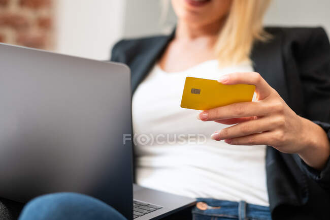 Cortado irreconocible rubia mujer en ropa casual entrar credenciales de tarjeta de crédito en el ordenador portátil mientras está sentado en un cómodo sillón y hacer compras en línea en casa - foto de stock