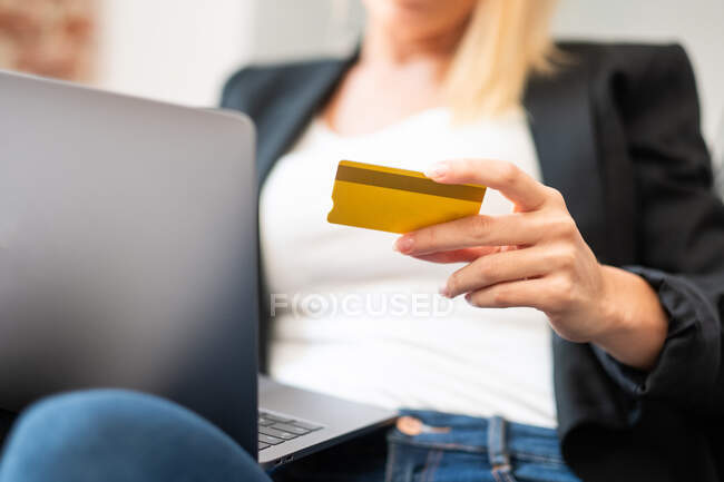 Cropped unrecognizable blonde female in casual clothes entering credit card credentials on laptop while sitting in comfortable armchair and making online purchases at home — Stock Photo
