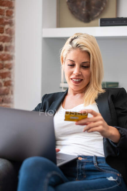 Blonde Frau in lässiger Kleidung gibt Kreditkartendaten auf Laptop ein, während sie im bequemen Sessel sitzt und zu Hause online einkauft — Stockfoto
