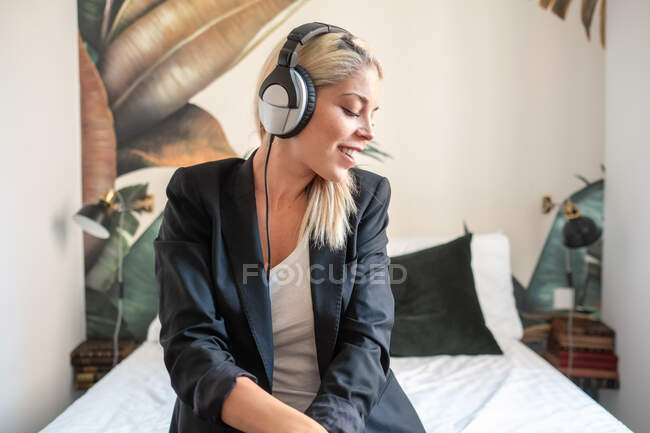 Glückliche moderne Frau mit Kopfhörern, die mit geschlossenen Augen lächelt und Musik hört, während sie zu Hause im Bett sitzt — Stockfoto