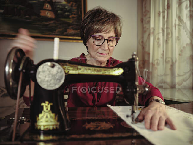 Старша леді в окулярах використовує ретро швейну машинку для створення лляної серветки в затишній кімнаті вдома — стокове фото