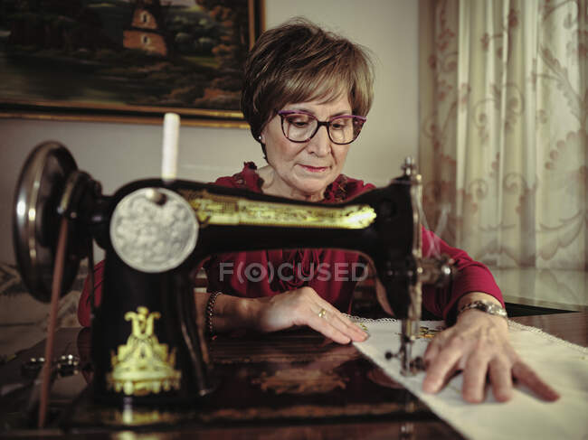 Señora mayor en gafas utilizando la máquina de coser retro para crear servilleta de lino en la habitación acogedora en casa - foto de stock