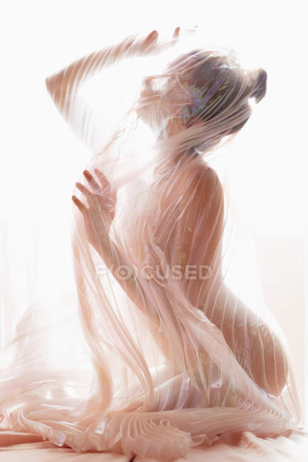 Вид збоку анонімної гола модель, вкрита прозорою плісированою тканиною завіси проти яскравого сонячного світла — стокове фото
