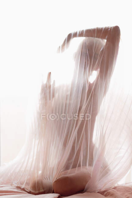 Анонимная обнаженная модель, покрытая прозрачной плиссированной тканью занавеса против яркого солнечного света — стоковое фото