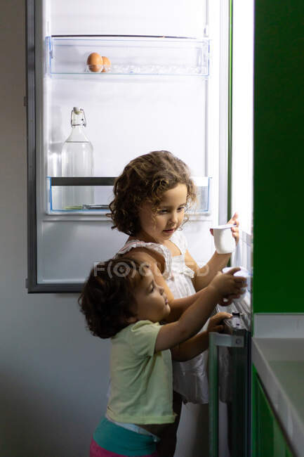 Little girl and toddler taking fresh yoghurt from fridge while stealing food at night at home together — Stock Photo