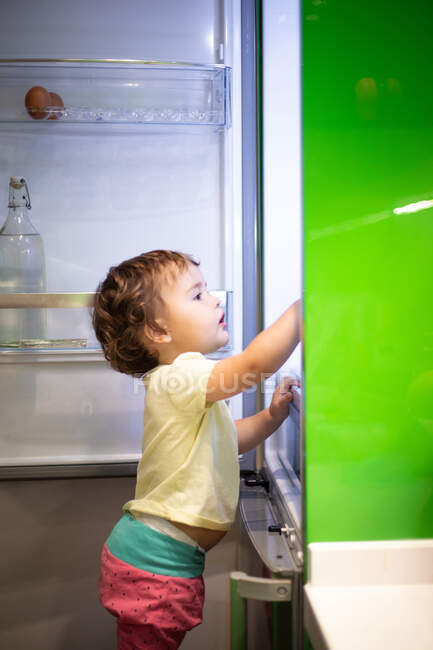 Seitenansicht des niedlichen kleinen Kindes, das auf einem Hocker steht und in der gemütlichen Küche zu Hause Lebensmittel aus dem offenen Kühlschrank nimmt — Stockfoto