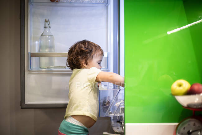 Vista lateral de un niño pequeño y lindo de pie en el taburete y tomar la comida del refrigerador abierto en la acogedora cocina en casa - foto de stock