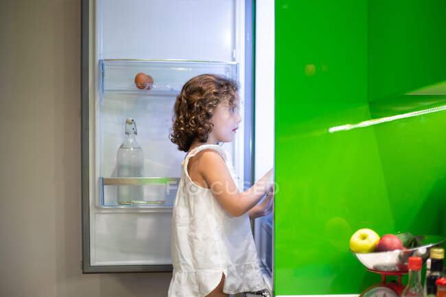 Menina em sleepwear à procura de lanche dentro da geladeira aberta à noite na cozinha em casa — Fotografia de Stock