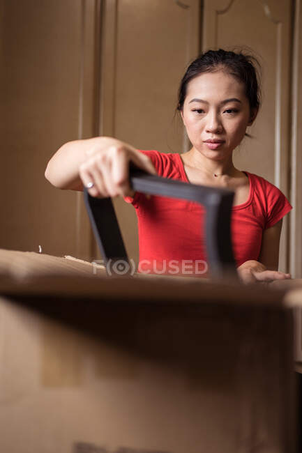 Ethnische Dame packt Karton mit zerlegten Stuhlteilen nach der Entbindung in gemütlichem Raum zu Hause aus — Stockfoto