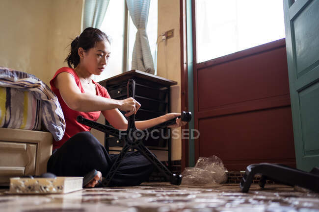 Seitenansicht der ethnischen asiatischen Frau Schrauben Räder auf Stuhluntergestell, während sitzen im Schneidersitz auf dem Boden in der Nähe Bett und Montage von Möbeln in gemütlichen Schlafzimmer zu Hause — Stockfoto