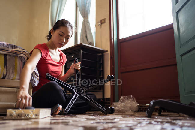 Vista lateral de las hembras asiáticas étnicas atornillando ruedas a la base de la silla mientras está sentado con las piernas cruzadas en el suelo cerca de la cama y el montaje de muebles en el acogedor dormitorio en casa - foto de stock