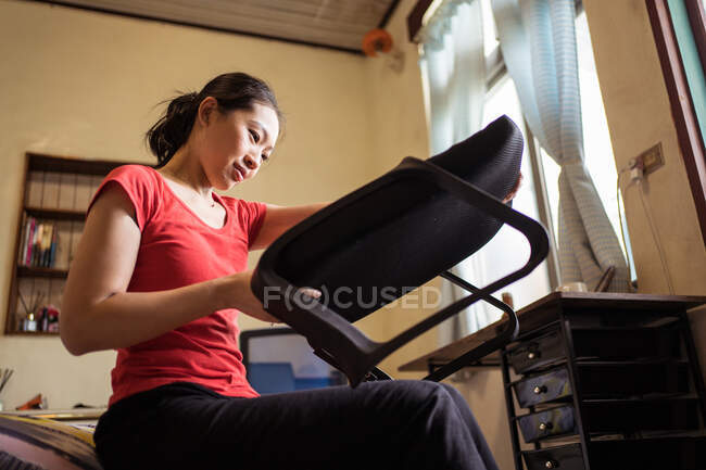Basso angolo di signora etnica seduta sul letto e controllo sedile morbido con maniglie durante il montaggio della sedia a casa — Foto stock