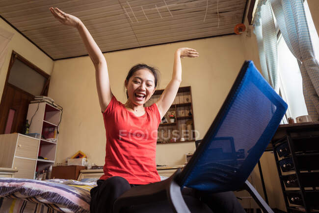 Basso angolo di eccitata donna asiatica con le braccia sollevate che celebrano il successo della sedia assemblare mentre seduto sul letto in camera da letto accogliente — Foto stock