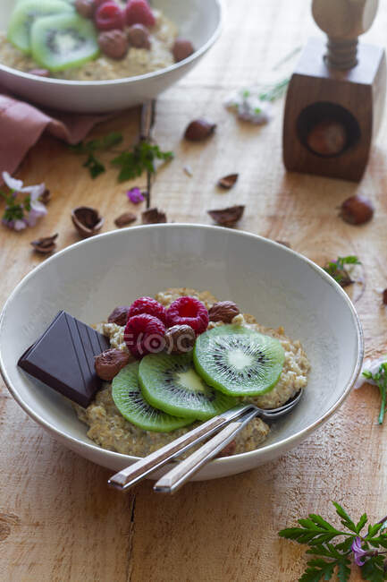De acima mencionadas fatias de kiwi fresco e framboesas colocadas perto de chocolate e avelãs em tigela de mingau de cereal saudável em mesa de madeira perto de pano lilás — Fotografia de Stock