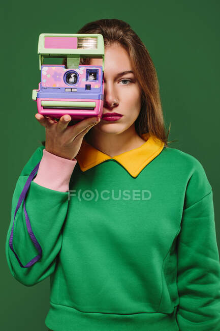 Молода неемоційна жінка в зеленому пуловері фотографує з ретро миттєвою камерою, стоячи на зеленому тлі — стокове фото