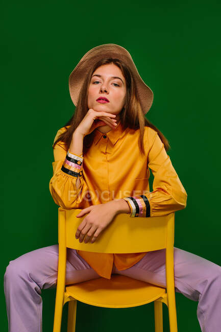 Trendy confident millennial female in colorful clothes and hat sitting on stool and looking at camera against green background — Stock Photo