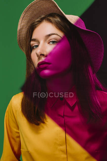 Young female model in stylish hat with red shadow on face and shoulder looking at camera against green background — Stock Photo