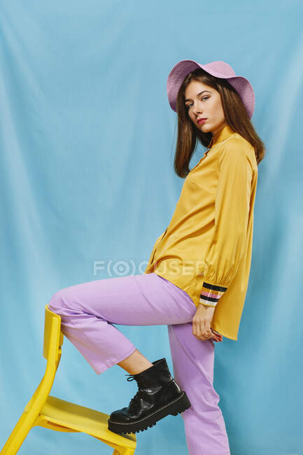 Seitenansicht der trendigen jungen selbstbewussten Frau in stilvoller farbenfroher Kleidung und Hut, die in die Kamera schaut, während sie auf einem gelben Stuhl vor blauem Hintergrund steht. — Stockfoto