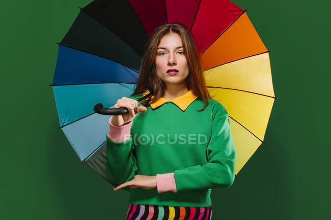 Young unemotional female model in colorful outfit holding multicolored umbrella and looking at camera while standing against green background — Stock Photo