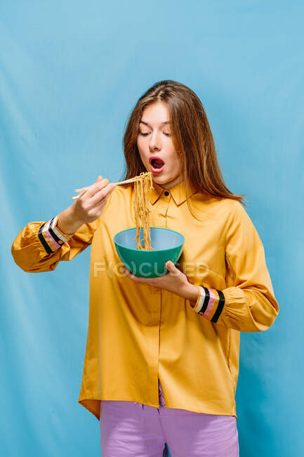 Giovane donna positiva in camicia gialla bocca di apertura per mangiare gustosi spaghetti istantanei con bacchette su sfondo blu — Foto stock