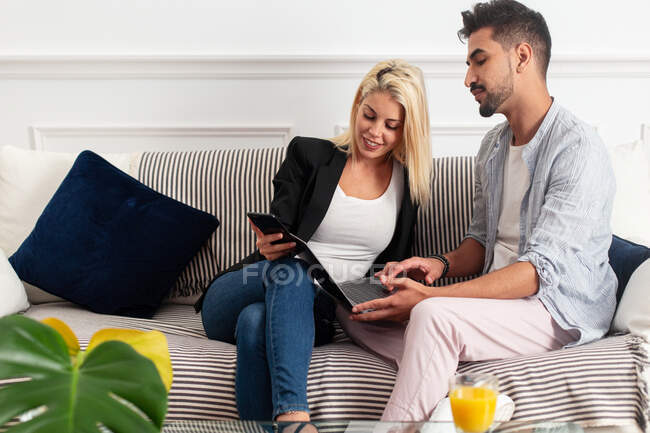 Mujer rubia positiva con taza de jugo de navegación teléfono inteligente y sentado en el sofá cerca de novio étnico escribiendo en el teclado del ordenador portátil en la sala de estar del apartamento moderno - foto de stock