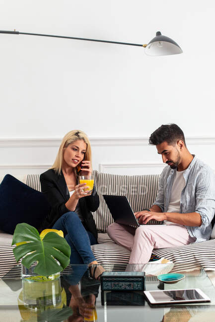 Позитивная блондинка с чашкой сока говорит на смартфоне и сидит на диване возле этнического бойфренда, печатая на клавиатуре ноутбука в гостиной современной квартиры — стоковое фото