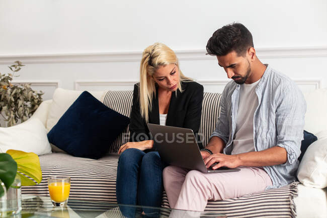 Donna bionda e barbuto etnico bell'uomo in abiti casual seduti sul divano e navigando laptop insieme a casa — Foto stock