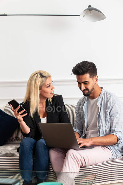 Mujer rubia positiva navegando por teléfono inteligente y sentado en el sofá cerca de novio étnico escribiendo en el teclado del ordenador portátil en la sala de estar del apartamento moderno - foto de stock