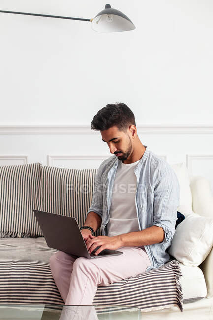 Молодой этнический человек в повседневной одежде сидит на диване и просматривает ноутбук в гостиной современной квартиры — стоковое фото