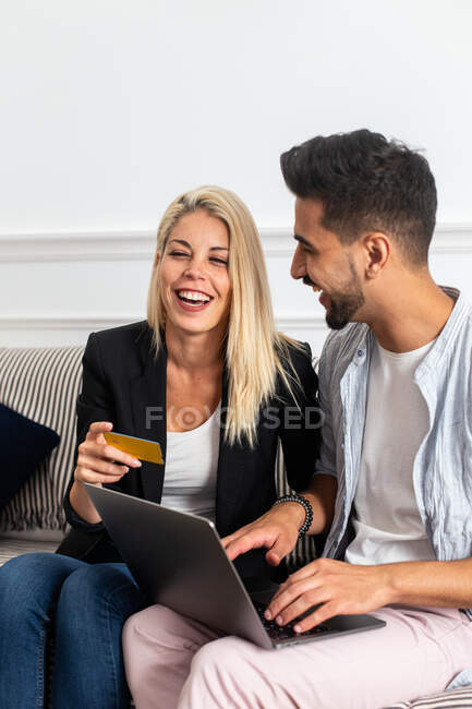 Deliciosa hembra rubia sonriendo y leyendo credenciales de tarjeta de crédito a alegre novio étnico con portátil mientras está sentado en el sofá y haciendo compras en línea juntos - foto de stock