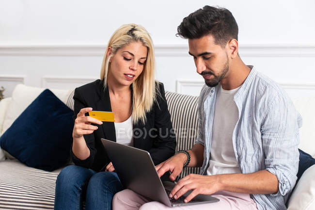 Deleitada hembra rubia sonriendo y leyendo credenciales de tarjeta de crédito a novio étnico con portátil mientras se sienta en el sofá y hacer compras en línea juntos - foto de stock