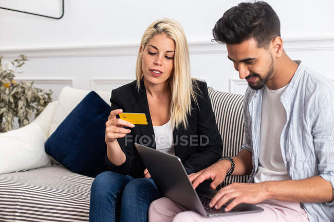 Deleitado hembra rubia sonriendo y leyendo credenciales de tarjeta de crédito a alegre novio étnico con portátil mientras se sienta en el sofá y hacer compras en línea juntos - foto de stock