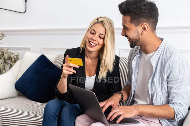 Délicieuse blonde femme souriante et lisant les informations d'identification de carte de crédit à petit ami ethnique joyeux avec ordinateur portable tout en étant assis sur le canapé et faire des achats en ligne ensemble — Photo de stock